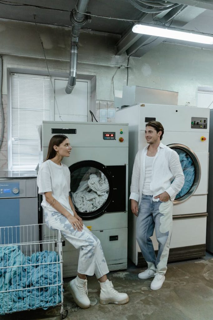 Two people in white clothing converse beside industrial laundry machines in a facility.